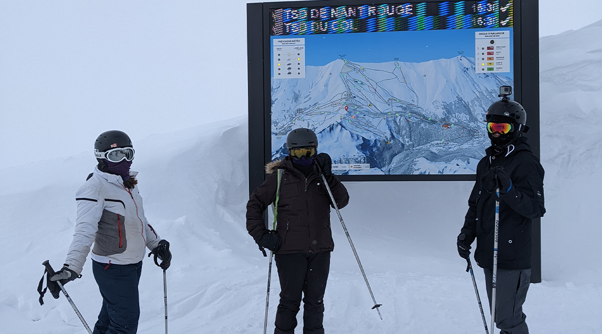 Three skiers stand by a big map board at the top of a chair lift